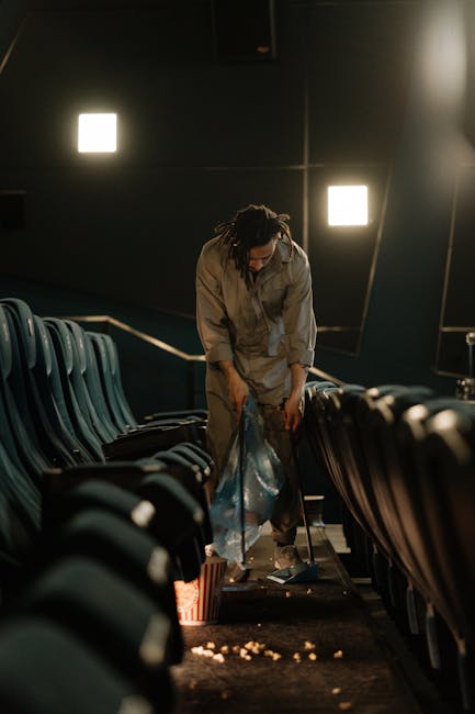 A professional cleaner from Earls Court Carpet Cleaning is performing deep cleaning or sanitisation in a darkened auditorium, with rows of blue upholstered chairs aligned on either side of a central aisle. The cleaner, dressed in a beige uniform, is using a vacuum or cleaning tool to remove debris from the carpeted floor, which shows scattered crumbs or dirt. Soft, square wall-mounted lights provide focused illumination in the room, highlighting the clean surfaces and the careful cleaning process. The setting emphasizes meticulous surface cleaning and hygienic maintenance, relevant to domestic and commercial cleaning services offered by Earls Court Carpet Cleaning.