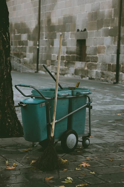 A green outdoor cleaning cart with a mop, broom, and other cleaning tools is positioned on a paved sidewalk, leaning against a tree. The cart contains cleaning supplies and is situated near a gray brick wall in an urban environment. Fallen leaves are scattered on the ground, and the scene appears to be in the daytime with natural lighting. This setup reflects typical equipment used in domestic and commercial surface cleaning, as highlighted by Earls Court Carpet Cleaning for their services related to avoiding hidden cleaning charges in Earls Court. The image emphasizes the importance of routine maintenance and cleanliness in public or outdoor areas, aligning with deep cleaning and sanitisation procedures.