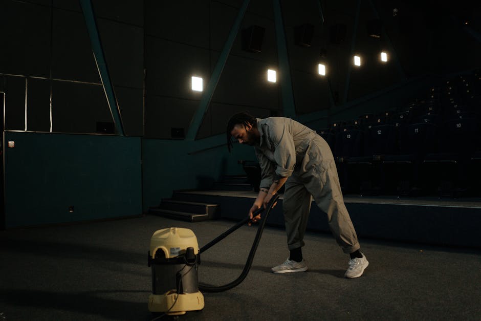A man performing surface cleaning in a large, indoor auditorium or cinema hall with dark walls and tiered seating. He is using a professional vacuum cleaner with a long hose attachment on the carpeted floor, wearing casual grey clothing and sneakers. The space is lit by overhead lights, creating a focused environment for deep cleaning and sanitisation. The area appears well-maintained and free of dust, with clean and vacuumed flooring, highlighting the cleaning process by Earls Court Carpet Cleaning for domestic and commercial spaces.