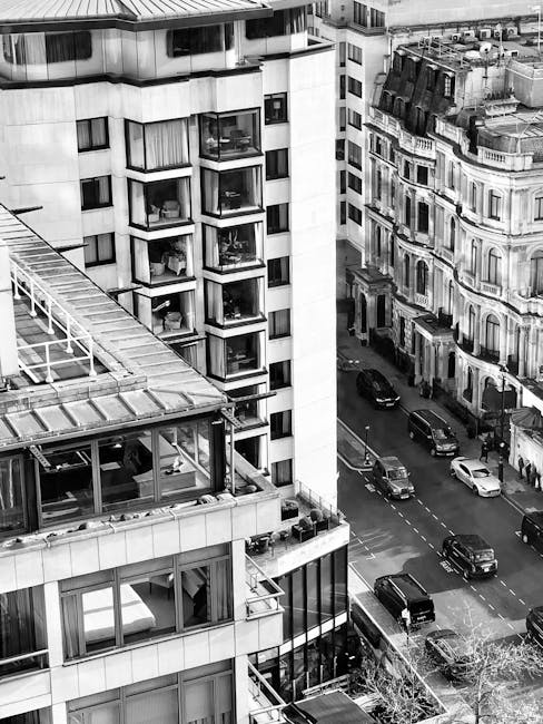 A black-and-white aerial view of a city street showing tall residential and commercial buildings with modern glass and concrete facades. The scene includes multiple levels of windows, some with visible balconies and interior furnishings, and street-level cars parked along the curb. The image captures the urban environment during daytime with natural lighting, emphasizing clean and well-maintained surfaces on the building exteriors. The scene reflects a busy cityscape typical of Earls Court, demonstrating the importance of professional surface cleaning and maintenance, as offered by Earls Court Carpet Cleaning, for residential and commercial properties.