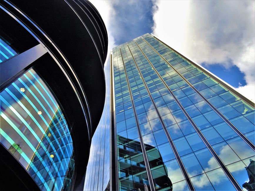 A modern office building with a glass exterior reflecting a partly cloudy sky, featuring vertical window panels and sleek metal framing. The building's facade is highly polished and clean, with visible interior lighting through the glass. The lower portion shows a curved section with blue lighting accents. The environment appears well-maintained, emphasizing the importance of surface cleaning and sanitisation, as promoted by Earls Court Carpet Cleaning on the Office cleaning Empress State Building Earls Court page.