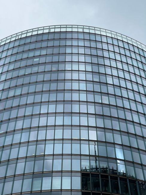 A modern, cylindrical office building with a curved glass facade featuring multiple horizontal rows of tinted windows. The reflective glass panels mirror the cloudy sky above, and the building's smooth, clean surface indicates regular surface cleaning and maintenance. The structure's height and design emphasize a sleek, contemporary architectural style, with no visible dirt or smudges on the glass. The image is taken from a low angle, highlighting the building's prominence and polished appearance, representative of professional cleaning services by Earls Court Carpet Cleaning for commercial properties like the Empress State Building in Earls Court.