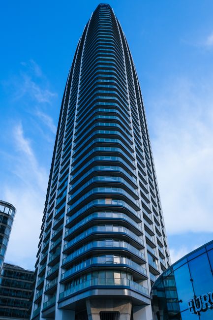 A modern office building with a glass exterior reflecting a partly cloudy sky, featuring vertical window panels and sleek metal framing. The building's facade is highly polished and clean, with visible interior lighting through the glass. The lower portion shows a curved section with blue lighting accents. The environment appears well-maintained, emphasizing the importance of surface cleaning and sanitisation, as promoted by Earls Court Carpet Cleaning on the Office cleaning Empress State Building Earls Court page.