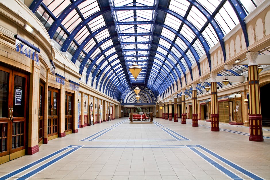A spacious indoor shopping arcade featuring a high, arched glass ceiling supported by white columns with decorative gold and red accents. The polished tiled floor is clean and shiny with a geometric pattern in blue and beige, reflecting the overhead lighting. Along the sides, there are multiple closed retail store entrances with wooden framed glass doors and signs, indicating a commercial setting. The area appears bright, well-maintained, and free of dust or debris, illustrating a professional hygienic environment suitable for surface cleaning and maintenance, as promoted by Earls Court Carpet Cleaning under their same day carpet cleaning services.