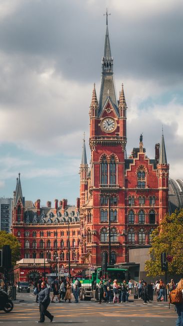 A busy urban scene featuring a large, historic red-brick building with ornate Gothic architectural details, including a tall clock tower topped with a spire. The foreground shows pedestrians walking on the street, some crossing at a crosswalk, and vehicles such as buses and cars driving past. The sky is partly cloudy with patches of blue. The image showcases the vibrant, bustling atmosphere of a city center, with visible greenery from trees lining the street. Earls Court Carpet Cleaning emphasizes surface cleaning and deep cleaning solutions suitable for such high-traffic areas to maintain hygiene and cleanliness, reflecting the company's expertise in sanitisation and domestic cleaning services.